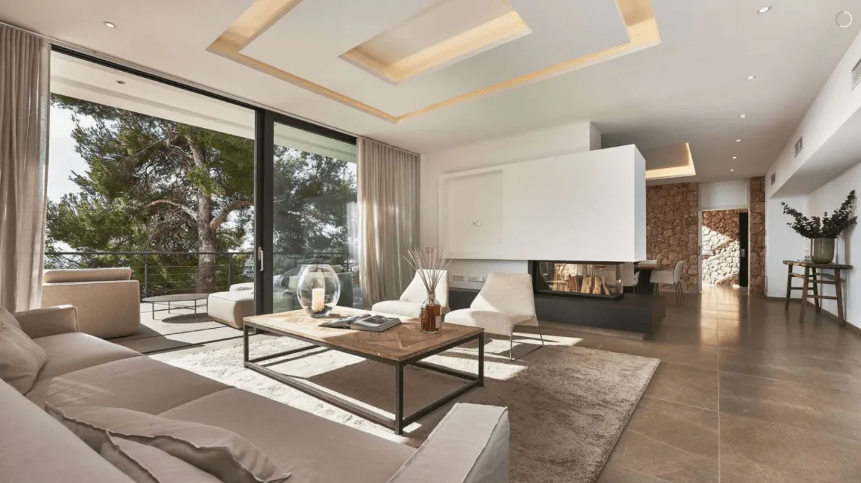 Salón de la vivienda Génova en Palma con chimenea central, grandes ventanales y pinos al exterior