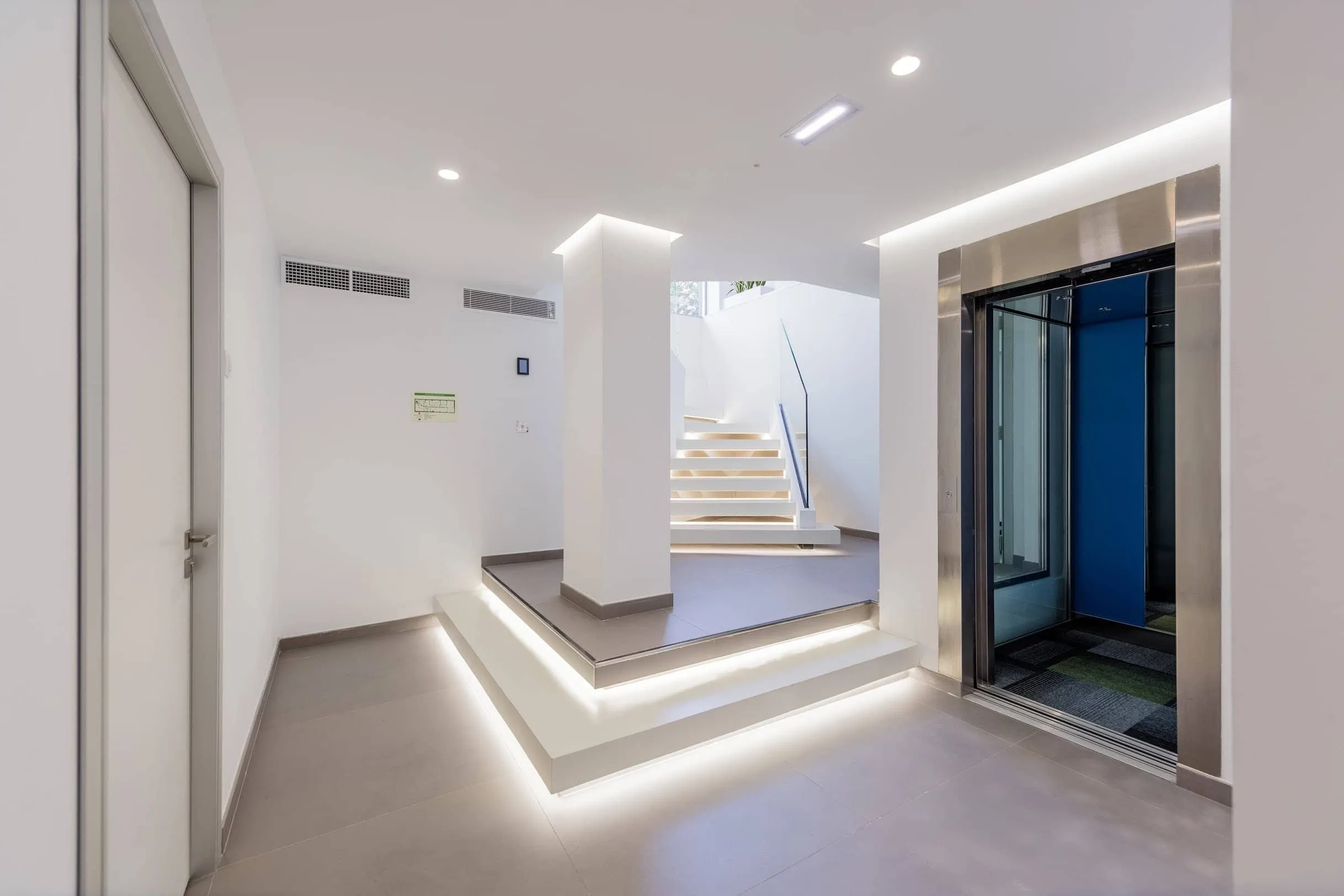 Descansillo con escalera blanca iluminada y ascensor en la reforma de oficinas SATSE, Palma